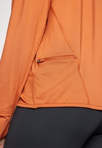 Oranžová ľahká športová top s bočnou zipsovou kapsou, plochými švami a priliehavým strihom. Látka vyzerá hladko a elasticky.
