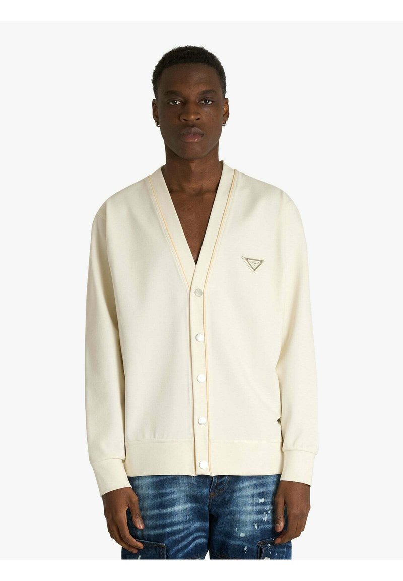 Cardigan crema chiaro con scollo a V profondo, bottoni bianchi e cuciture arancioni sottili. Indossato sopra jeans blu strappati.