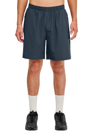 Mężczyzna w ciemnoszarych krótkich spodenkach sportowych, białych skarpetach do kostek i czarnych butach sportowych, stojący z nogami rozstawionymi na szerokość barków.