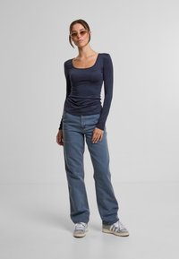 Granatowa bluzka z długim rękawem i okrągłym dekoltem, zestawiona z jasnoniebieskimi dżinsami o prostym kroju oraz białoszarymi sneakersami, podkreślona cienkimi okularami przeciwsłonecznymi.