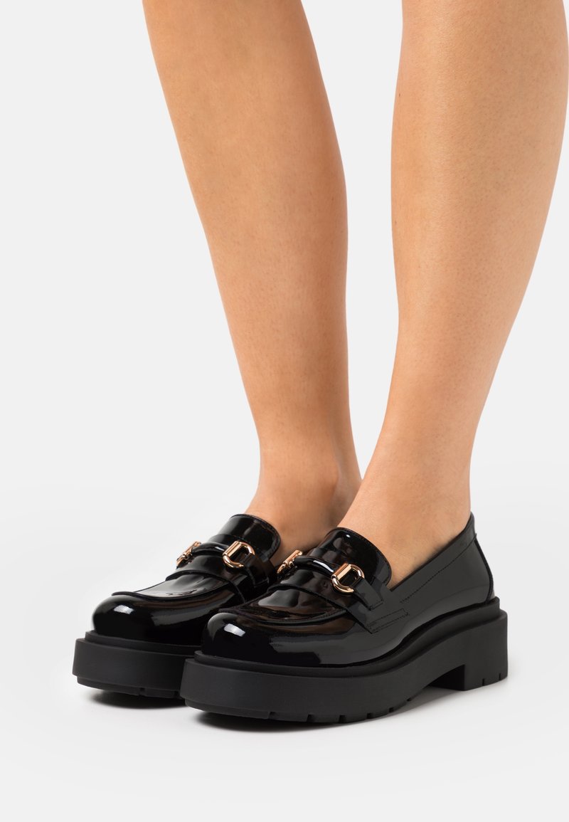 Mocassins en cuir verni noir avec quincaillerie dorée, semelles épaisses en plateforme noire et design de bout arrondi. Texture lisse avec une finition brillante.