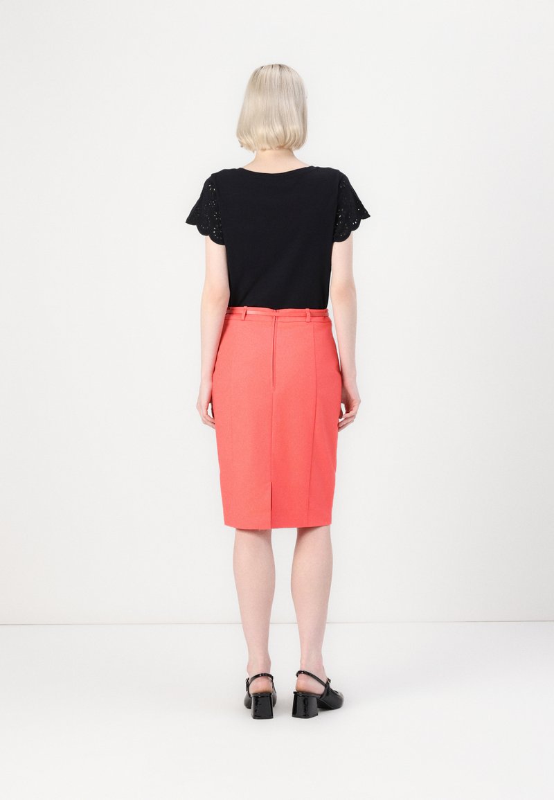 T-shirt noir avec des manches en dentelle, associée à une jupe crayon corail avec une fente à l'arrière, et des chaussures à talons carrés en vernis noir.