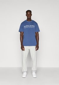 Modré bavlněné tričko s bílým textem "BJÖRN BORG TRAINING CLUB MONACO" v kombinaci s světle šedými tepláky a bílými teniskami. Jednoduchý, neformální design.