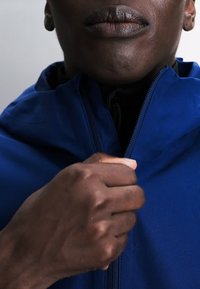 Gros plan sur la main d’une personne tirant la fermeture éclair d’une veste bleue, mettant l’accent sur les lèvres, le menton et les détails de la main.