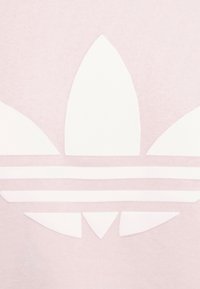 Gaiši rozā T-krekls ar lielu baltu Adidas trefoil logo un trim horizontālām līnijām zem tā. Gluda auduma tekstūra.