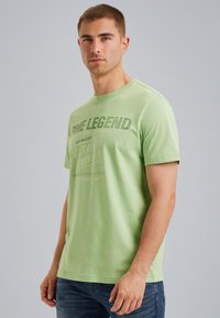T-shirt en coton vert clair avec des manches courtes. Le devant présente un texte graphique en vert foncé. Encolure ronde, coupe décontractée. Porté avec un jean bleu.