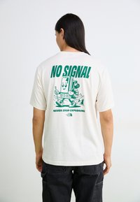 Cremefarvet bomulds T-shirt med et grønt grafik på ryggen af en smartphone-figur med teksten "INGEN SIGNAL" og "STOP ALDRIG MED AT UDFORSKE."