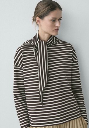 Frau mit braun-weiß gestreiftem Langarmshirt mit schalähnlichem Kragen, blickt seitlich vor schlichtem Hintergrund.