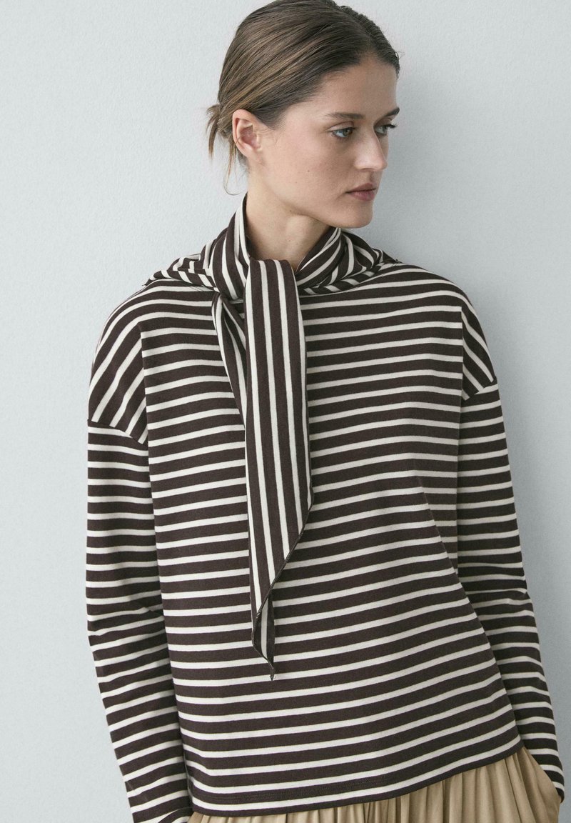 Kvinde iført en brun- og hvidstribet trøje med lange ærmer og et tørklæde-lignende halsdetalje, der kigger til siden mod en ensfarvet baggrund.