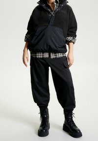 Schwarzer Fleece-Pullover mit hohem Kragen und Knopfleiste, darüber getragen ein schwarz-weiß kariertes Hemd. Kombiniert mit schwarzen Cargohosen und Stiefeln.