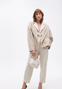 En modell poserar självsäkert i en skräddarsydd beige jacka och matchande byxor, medan hon håller en stilren vit handväska med en guldkedja.