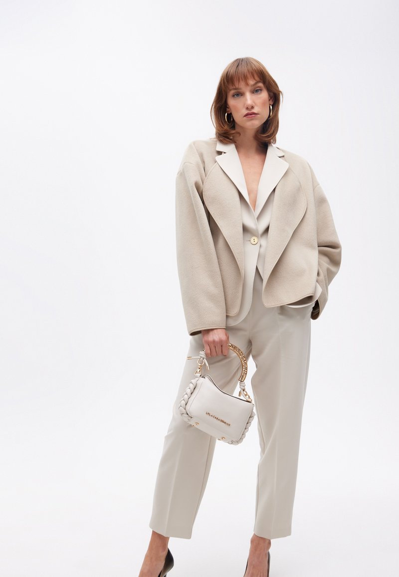 En modell poserar självsäkert i en skräddarsydd beige jacka och matchande byxor, medan hon håller en stilren vit handväska med en guldkedja.