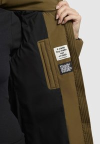 Olive green jacket interior with black lining, featuring two labels: one with text and another with a logo. Distinct quilted stitching visible.