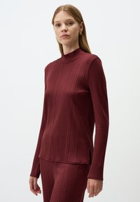 Bordeauxfarbener, gerippter Rollkragenpullover mit langen Ärmeln, der einen hohen Kragen und subtile seitliche Schlitze für optimale Bewegungsfreiheit bietet. Weiche Textur.