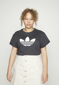 Juodas medvilnės marškinėliai trumpomis rankovėmis su dideliu baltu Adidas trefoil logotipu, apvaliu kaklu, derinami su šviesios spalvos marškinių sijonu.