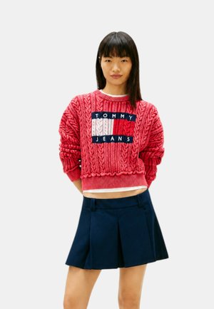 Junge Frau trägt einen roten Tommy Jeans Zopfpullover und einen dunkelblauen plissierten Rock und steht mit den Händen hinter dem Rücken.
