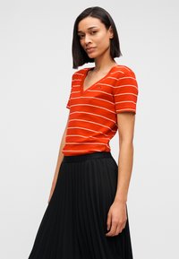 Κόκκινη μπλούζα με λαιμόκοψη σε σχήμα V με λευκές ρίγες, κοντά μανίκια και λεπτομέρεια λογότυπου. Συνδυασμένη με μαύρη φούστα με πιέτες, λεία υφή και ρέουσα σχεδίαση.