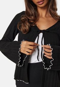 Cardigan nero a coste con maniche svasate e rifiniture in raso bianco, dotato di chiusura con fiocco davanti. Morbido al tatto e dal design aderente.