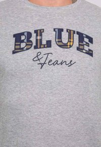 T-shirt gris à manches courtes présentant les inscriptions "BLUE" et "Jeans" en lettrage brodé. Les lettres mélangent des motifs unis et à carreaux en bleu et jaune.