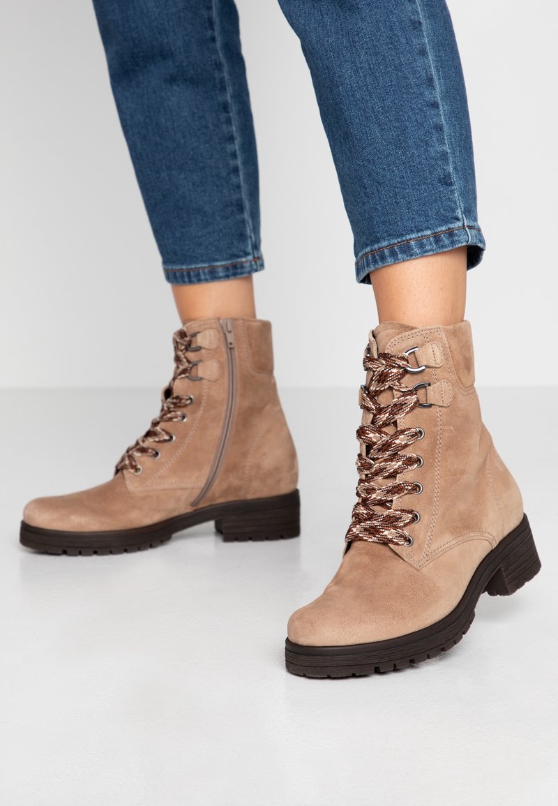 Bottines beiges en daim à lacets avec fermeture éclair latérale portées avec un jean bleu retroussé sur un sol blanc.