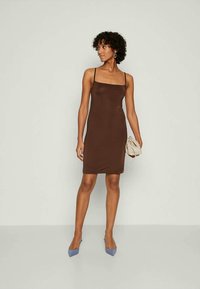 Nő, aki ujjatlan, barna, térd középig érő ruhát visel, egy kis bézs táskát tart a kezében, göndör haja van és kék hegyes orrú magassarkút visel.