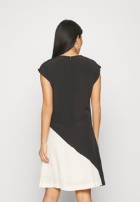 Kurzes Kleid mit einem schwarzen Oberteil und einem cremefarbenen, diagonal geschnittenen Unterteil. Kurze Ärmel und ein Reißverschluss im Rücken. Glattes Material mit leichtem Glanz.
