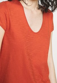 Kvinde iført en kortærmet, dyb scoop-neck orange skjorte med krøllet mørkt hår synligt på en hvid baggrund.