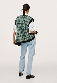 Grüne karierte Weste mit schwarzem Besatz, über einem weißen Hemd getragen, kombiniert mit hellblauen, zerrissenen Jeans und schwarzen Schuhen. Glatte Stofftextur.