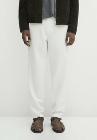 Pantaloni bianchi, giacca scura in suede e sandali aperti marroni. L'outfit presenta un maglione grigio a trama sotto la giacca.