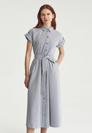 Joven mujer con un vestido camisero midi de rayas en azul claro y blanco, con botones y cinturón anudado en la cintura, de pie con una mano en el bolsillo.