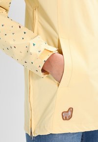 Hand i fickan på en blekgul jacka med ett litet djurmärke nära fållen och ärm med hjärtmotiv och kardborrestängning.