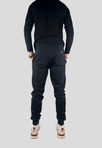 Černé sportovní tepláky s elastickým pasem, bočními kapsami a malým logem. Na nohou bílé a červené tenisky, zobrazené zezadu.