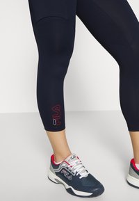 Granatowe legginsy o dopasowanym kroju z czerwonym logo Fila. Połączone z granatowo-białymi butami sportowymi z czerwonymi akcentami i perforowanymi detalami.