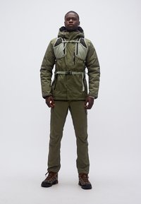 Άνδρας που στέκεται φορώντας εξωτερικό μπουφάν ελιάς πράσινου χρώματος, παντελόνια που ταιριάζουν, ορειβατικά μποτάκια και ιμάντα σακιδίου ορειβασίας σε απλό φόντο.