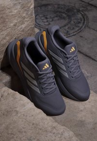 Paire de chaussures de course grises avec des rayures blanches et des accents jaunes placées sur des marches en pierre et un trottoir.