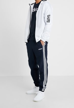 Persona con pantalones deportivos Adidas azul marino con rayas blancas, camiseta negra Adidas, chaqueta blanca con rayas negras en las mangas y zapatillas blancas.