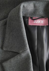 Manteau en mélange de laine gris avec un col cranté, doté d'une doublure noire lisse et d'une étiquette rose portant l'inscription "JJXX" cousue à l'intérieur.