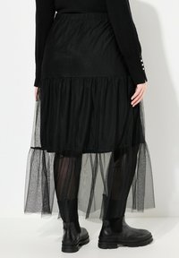 Schwarzer Maxirock mit geschichteter Tüllgestaltung, elastischem Bund und transparenter Textur. Tragen mit Knöchelstiefeln, die eine moderne Silhouette zeigen.
