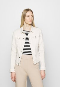 Giacca di jeans bianca con due tasche sul petto, bottoni argentati e una vestibilità strutturata, abbinata a una maglietta a righe bianco e nera e pantaloni beige.