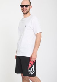 Homme souriant portant des lunettes de soleil noires, un t-shirt blanc et un short noir avec un design géométrique rouge et blanc sur le côté.