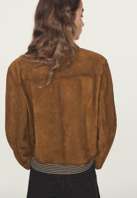 Persona con lunghi capelli mossi, vestita con una giacca in suede marrone e pantaloni scuri, vista di spalle su uno sfondo chiaro e semplice.