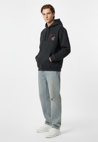 Felpa nera con tessuto testurizzato, polsini a costine e tasca a marsupio. Presenta un cuore rosso e un design grafico sul petto. Indossata con jeans azzurri chiari.