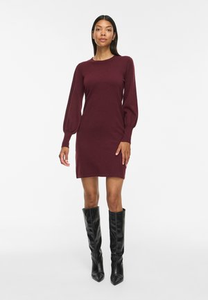 Femme portant une robe bordeaux ajustée à manches longues avec des manches bouffantes et des bottes noires à bout pointu montant jusqu’aux genoux, debout devant un fond blanc.