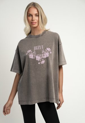 Blonde vrouw die een oversized vervaagd grijs T-shirt draagt met een roze bloemenpatroon en de tekst "BIZUU", gecombineerd met zwarte legging op een effen achtergrond.