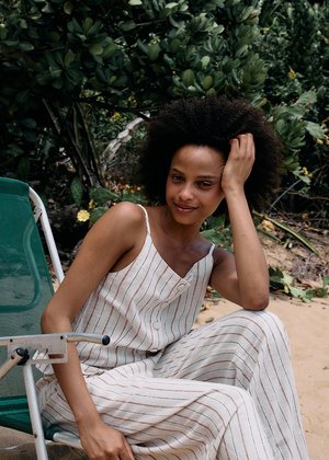 Göndör hajú nő csíkos ujjatlan overált visel, és kényelmesen ül egy zöld széken a szabadban, zöld növényzet és homokos talaj között.