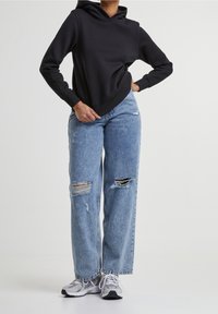 Czarna bluza z kapturem z kieszenią kangurką, zestawiona z jasnoniebieskimi, szerokimi spodniami typu jeans z przetarciami i dziurami. Do tego szare sneakersy.