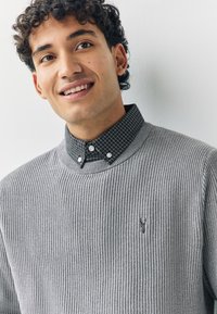 Grå ribbad tröja med rund halsringning, bärs över en svartvit rutmönstrad skjorta med knappar och detalj på kragen synliga.