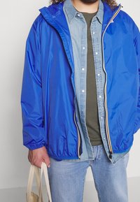 Blauwe waterdichte jas met elastische manchetten, gedragen over een denim overhemd en een olijfgroen T-shirt, gecombineerd met lichtblauwe spijkerbroek en een beige handtas.