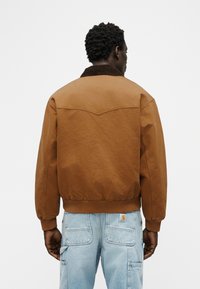 Άνδρας που φαίνεται από πίσω φοράει καφέ μπουφάν με σκούρο καφέ γιακά και ανοιχτό μπλε τζιν παντελόνι με πολλές τσέπες.