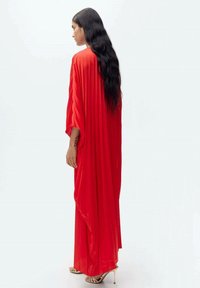 Kaftan merah yang mengalir dengan lengan panjang, lipatan vertikal, dan potongan longgar. Dipakai di atas celana lebar, dipadukan dengan sepatu heels bertali.